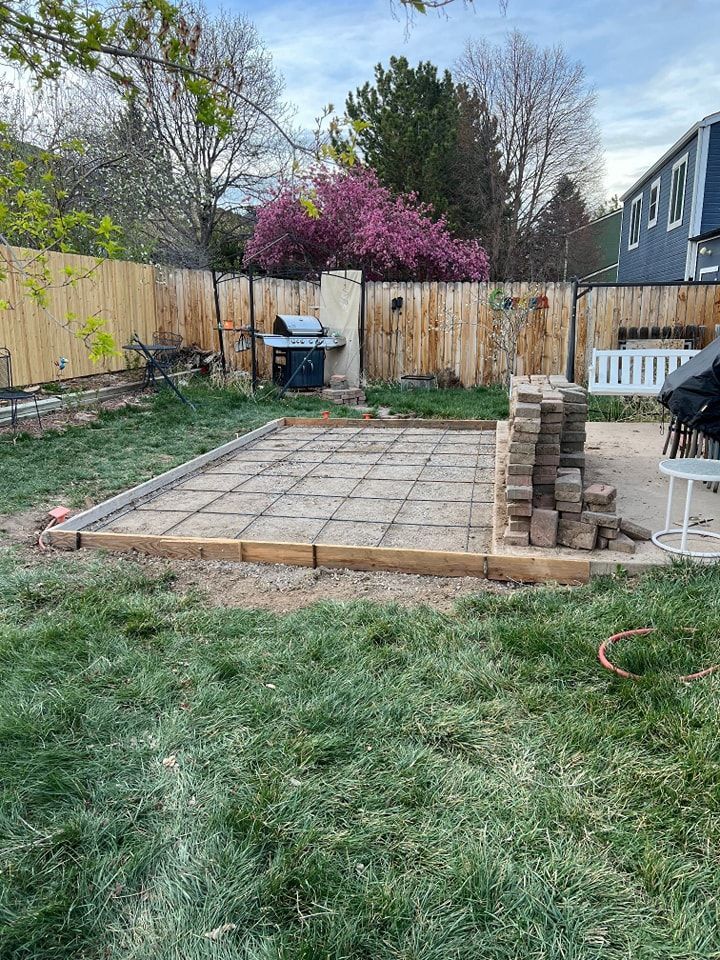 A backyard patio construction site with a square grid of pavers laid inside a wooden frame, with a stack of bricks nearby.