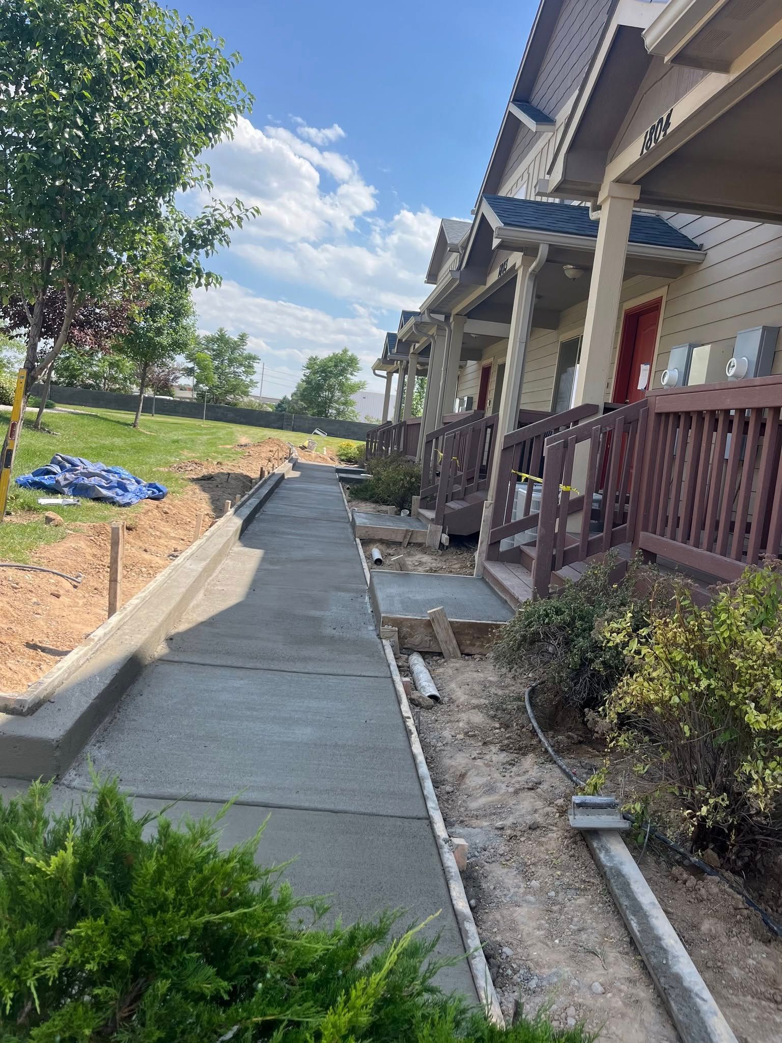 A sidewalk under construction runs beside a row of houses with brown wooden porches under a blue sky with white clouds.