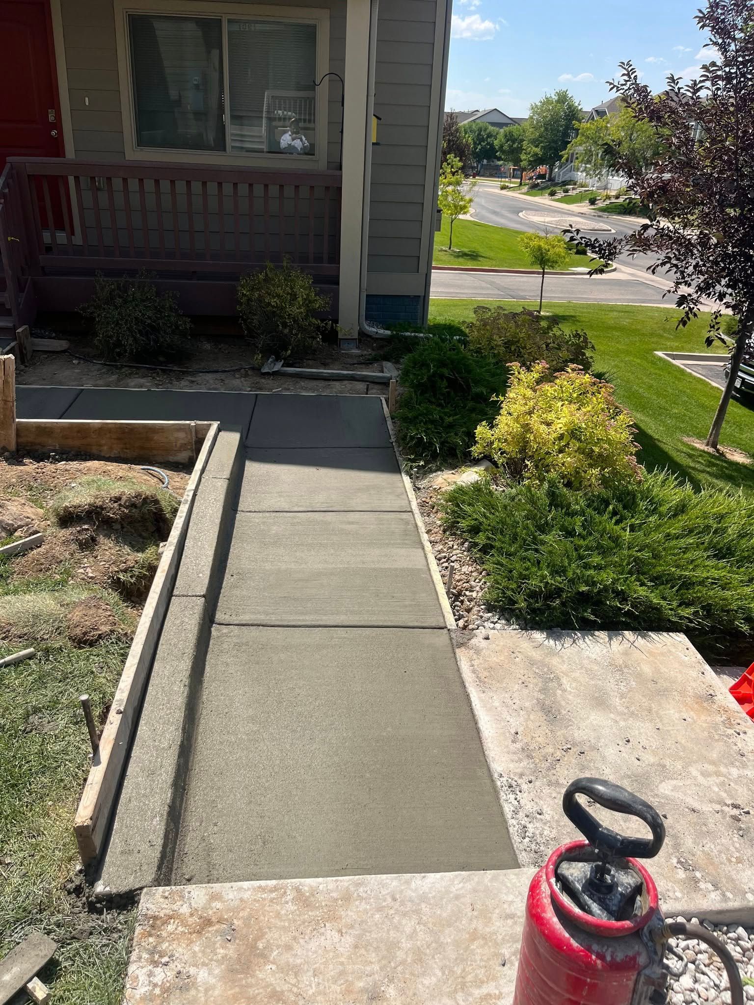 A newly poured concrete walkway leads to the front porch of a house, framed by wooden forms and landscaping.