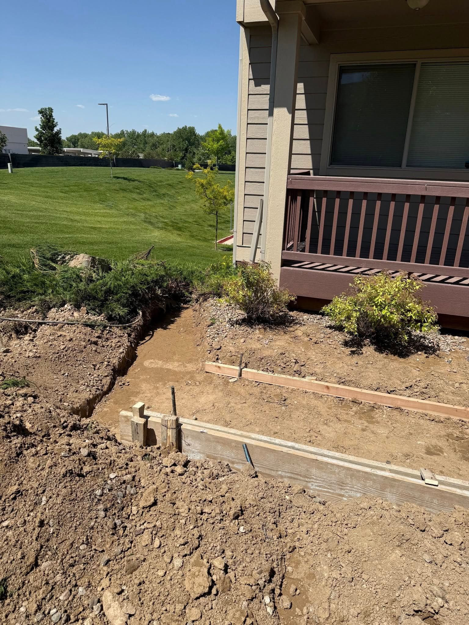 A construction site next to a house, showing a trench and wooden forms set in the ground for a concrete walkway.