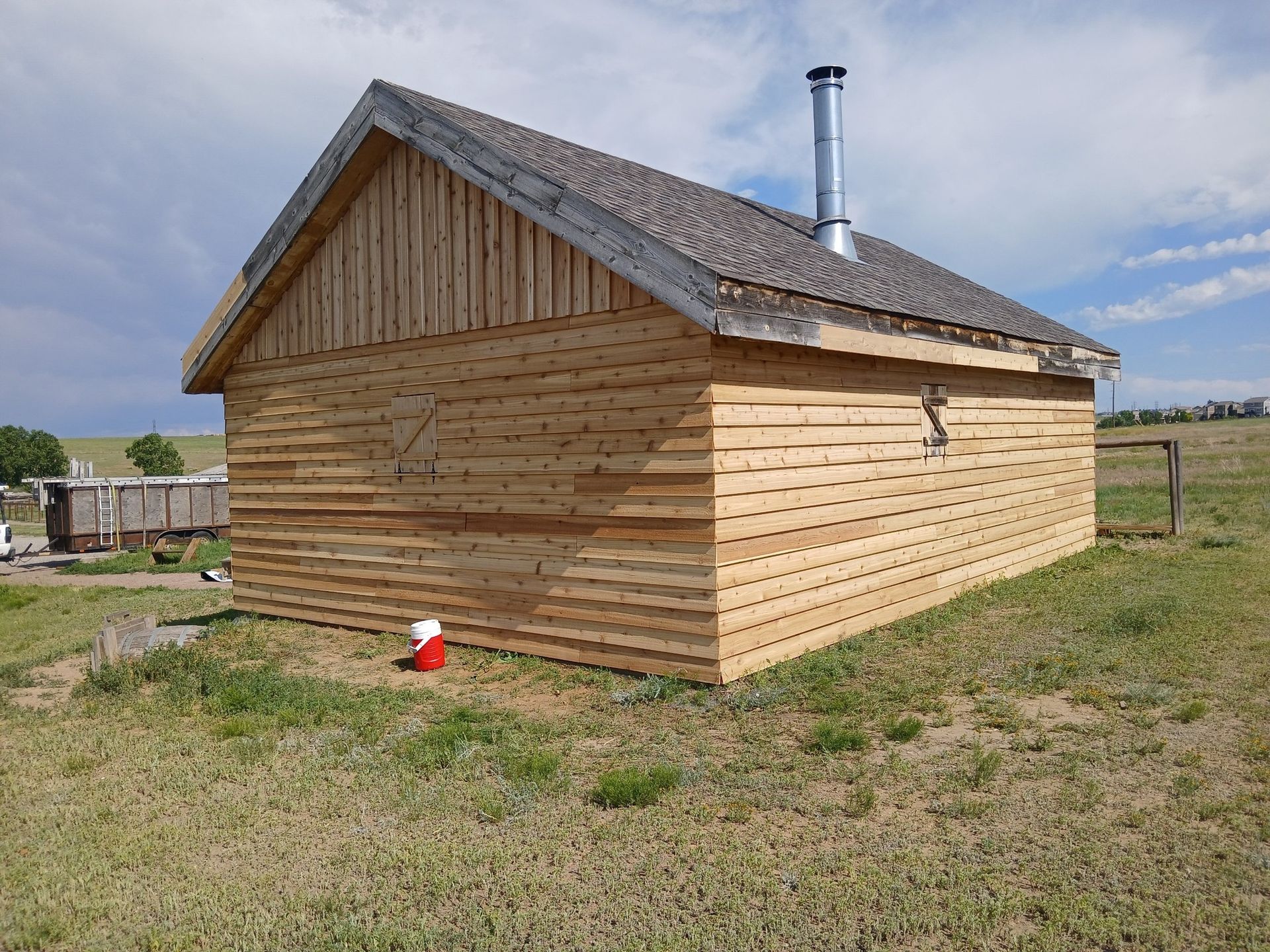 A small, wooden shed with horizontal siding and a metal chimney pipe on its shingled roof, standing in an open grass field.