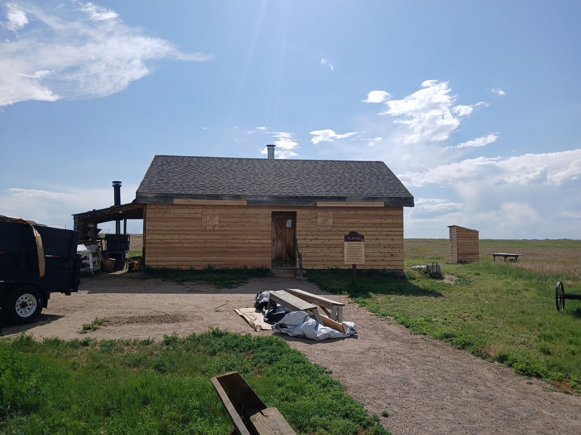 A small wooden cabin with horizontal siding and a metal chimney stands in a grassy, open field under a blue sky.