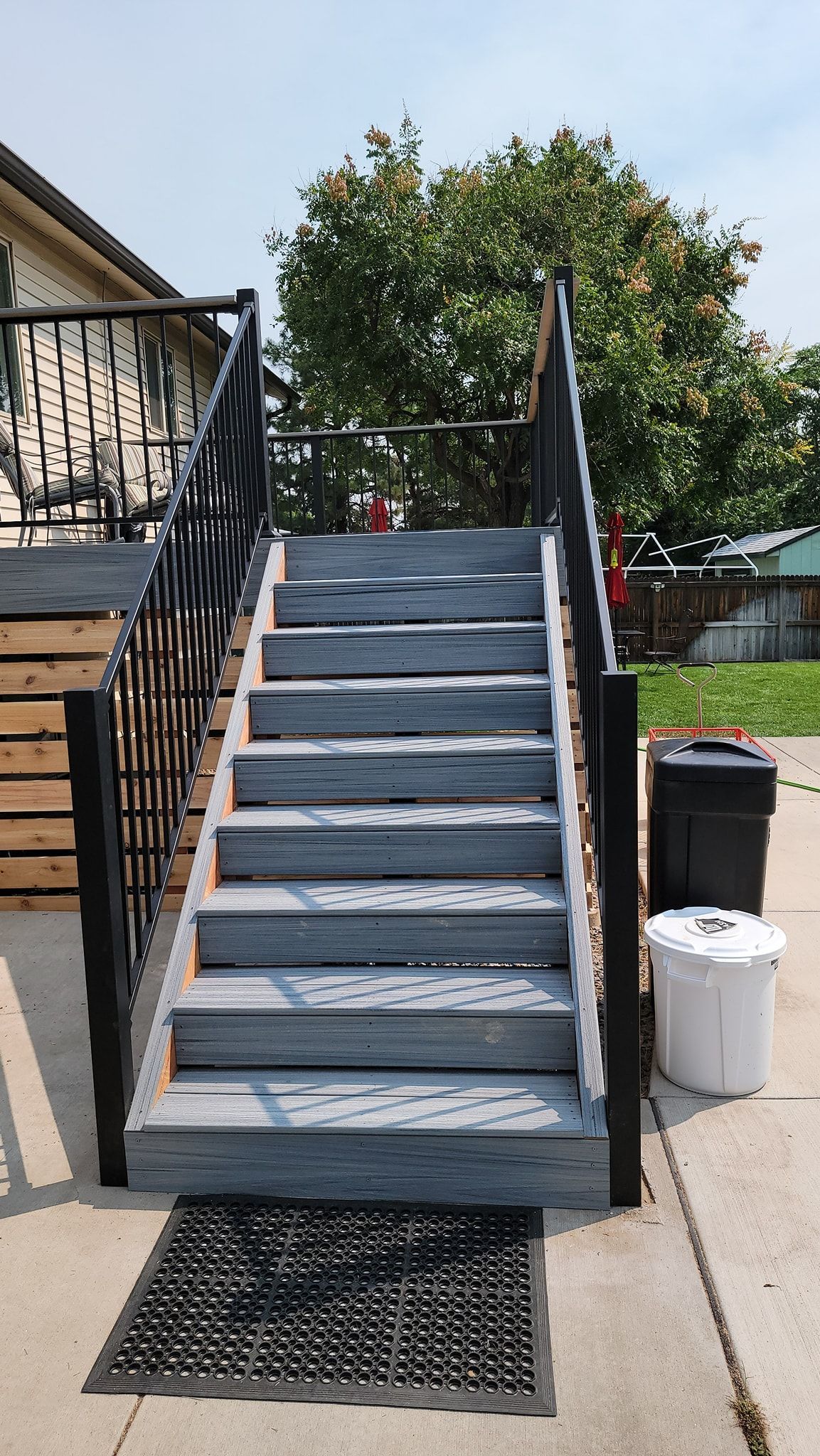 A set of grey wooden stairs with black railings leading up to a building entrance, with a mat and bins on the concrete.