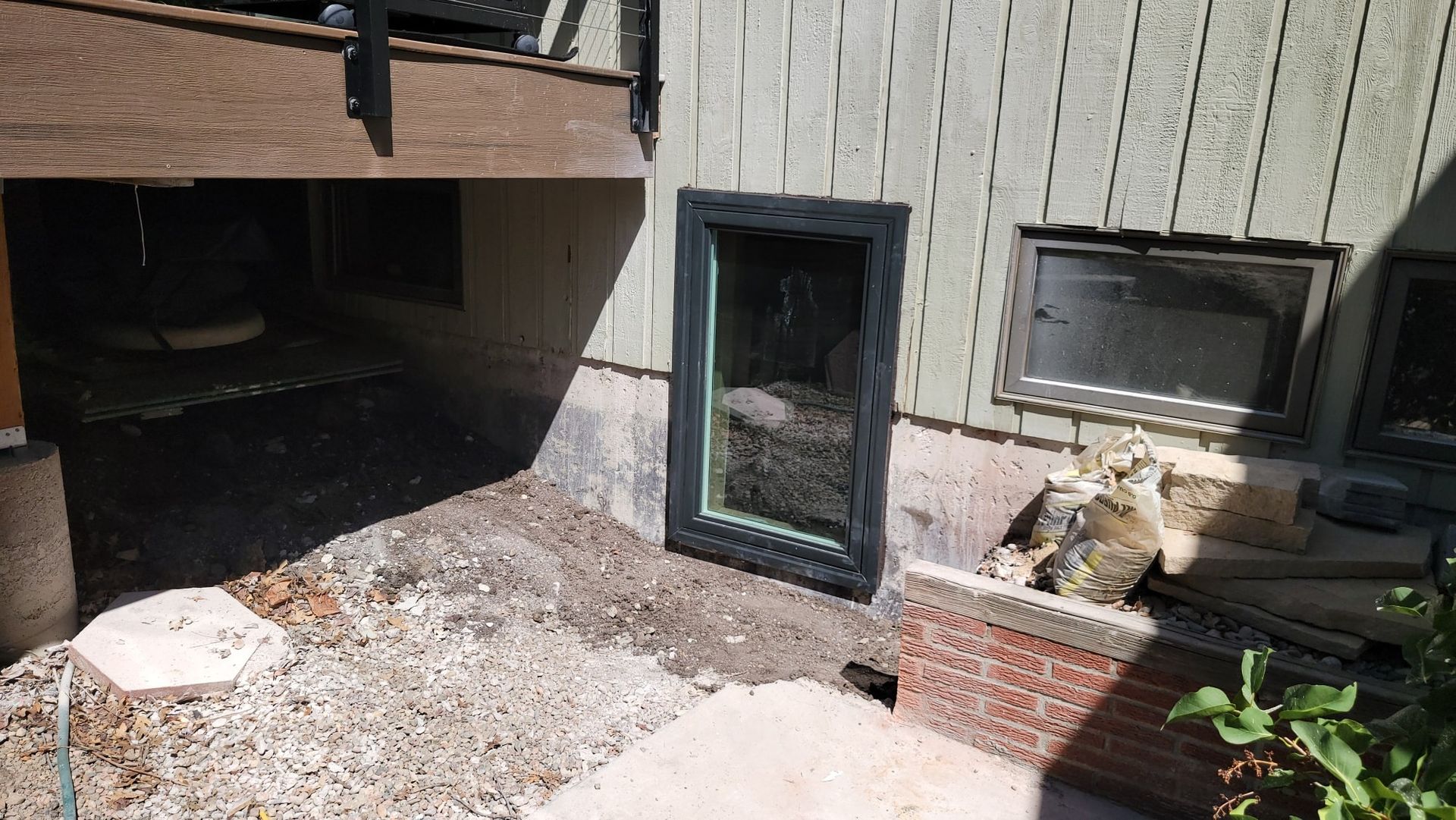 A newly installed tall, black-framed basement window on a beige siding wall next to an existing small, rectangular window.