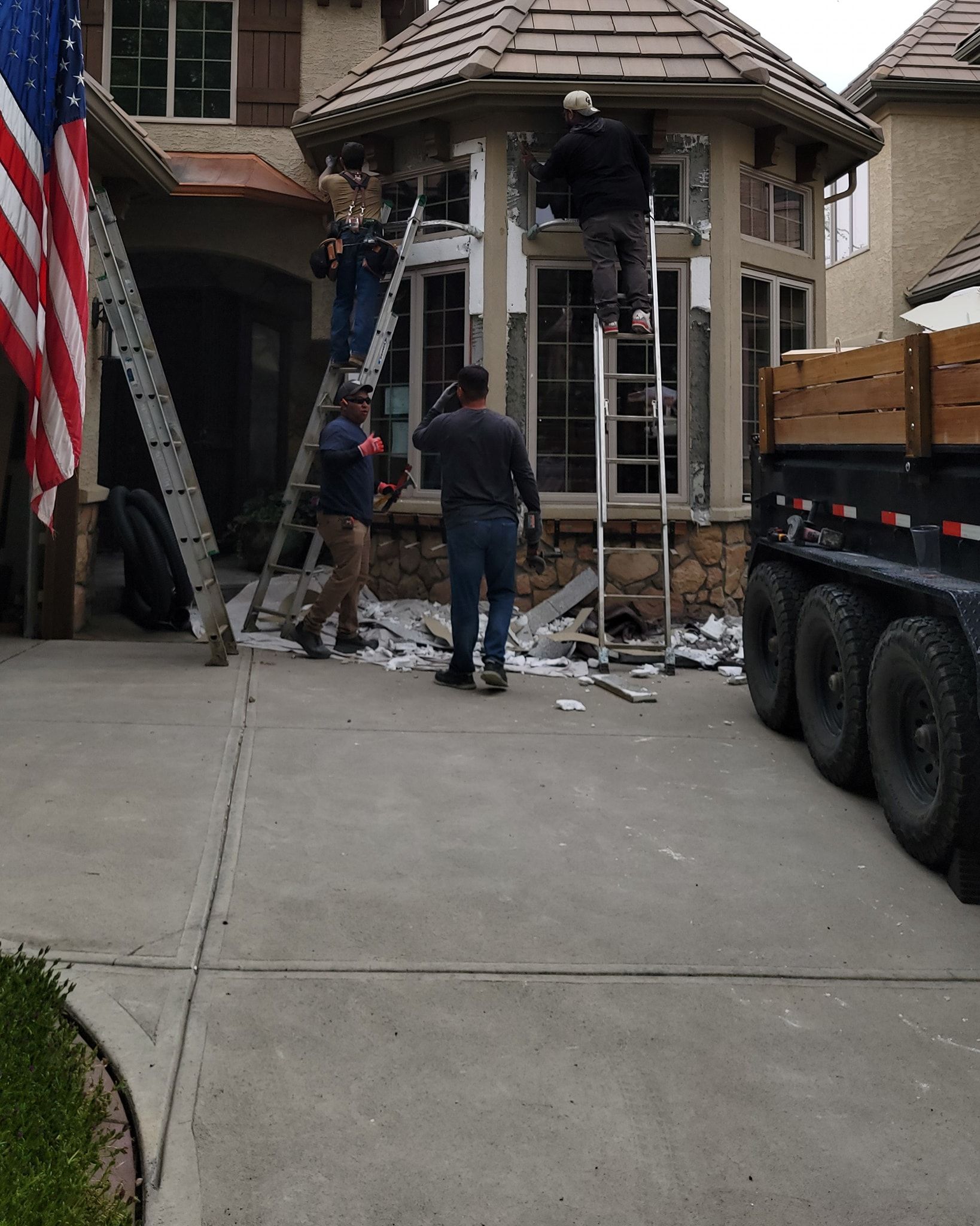 Workers in dark clothing perform exterior home renovations on a tan house with a dumpster parked nearby.