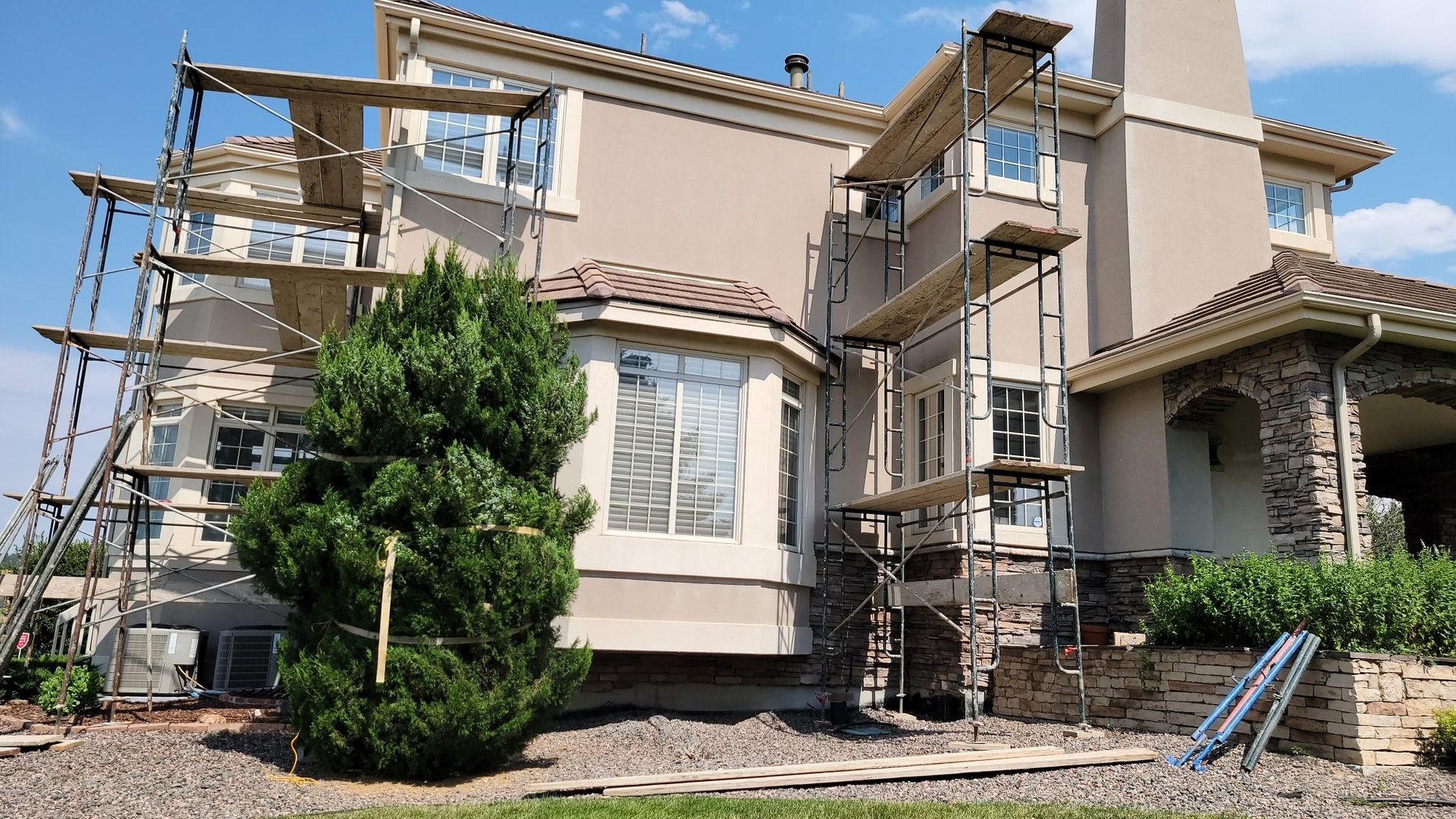 Scaffolding surrounds a two-story beige house with stone accents and a tiled roof, indicating exterior renovations.