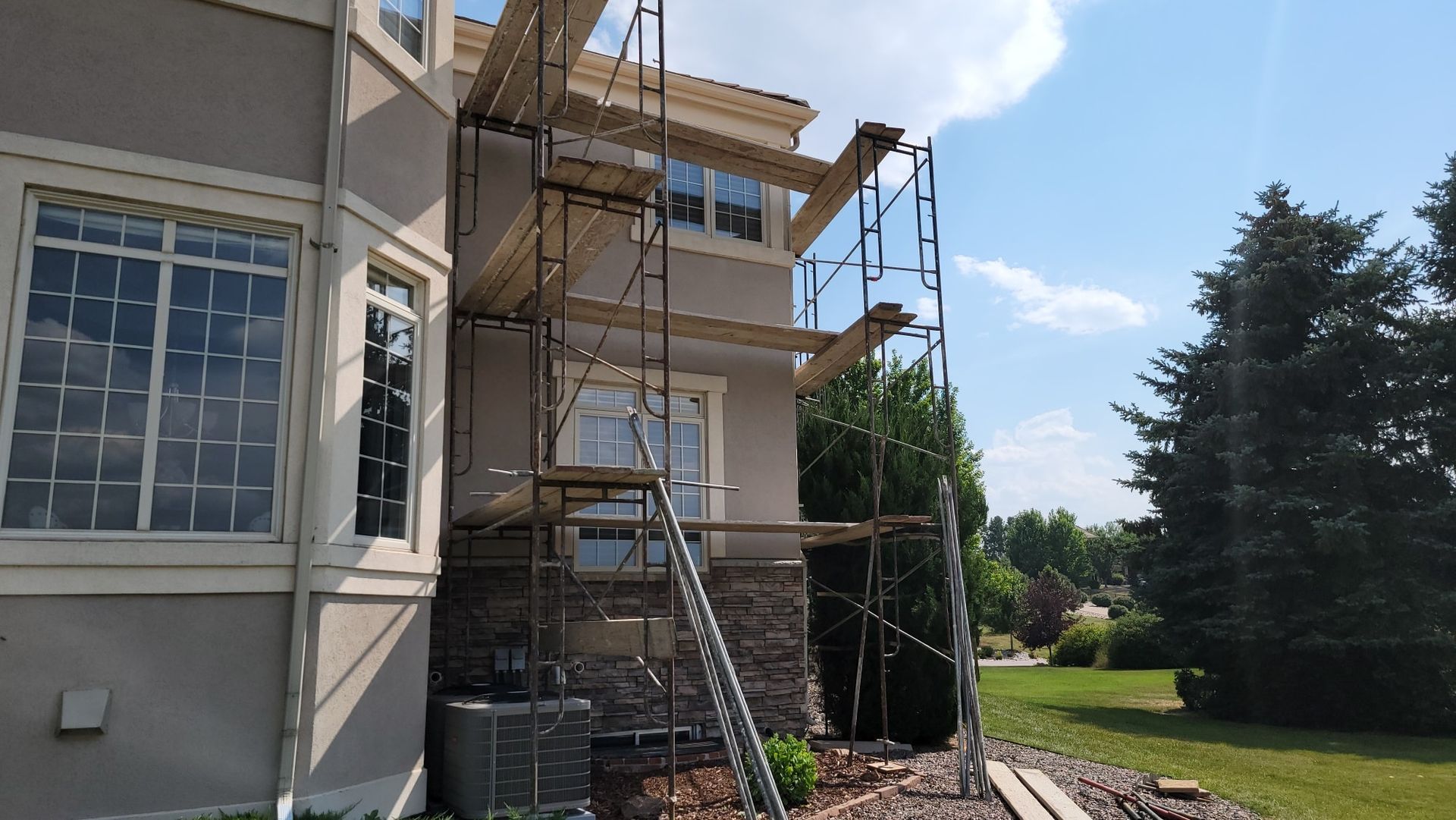 A two-story house exterior under construction, featuring metal scaffolding placed against a stone and stucco wall.