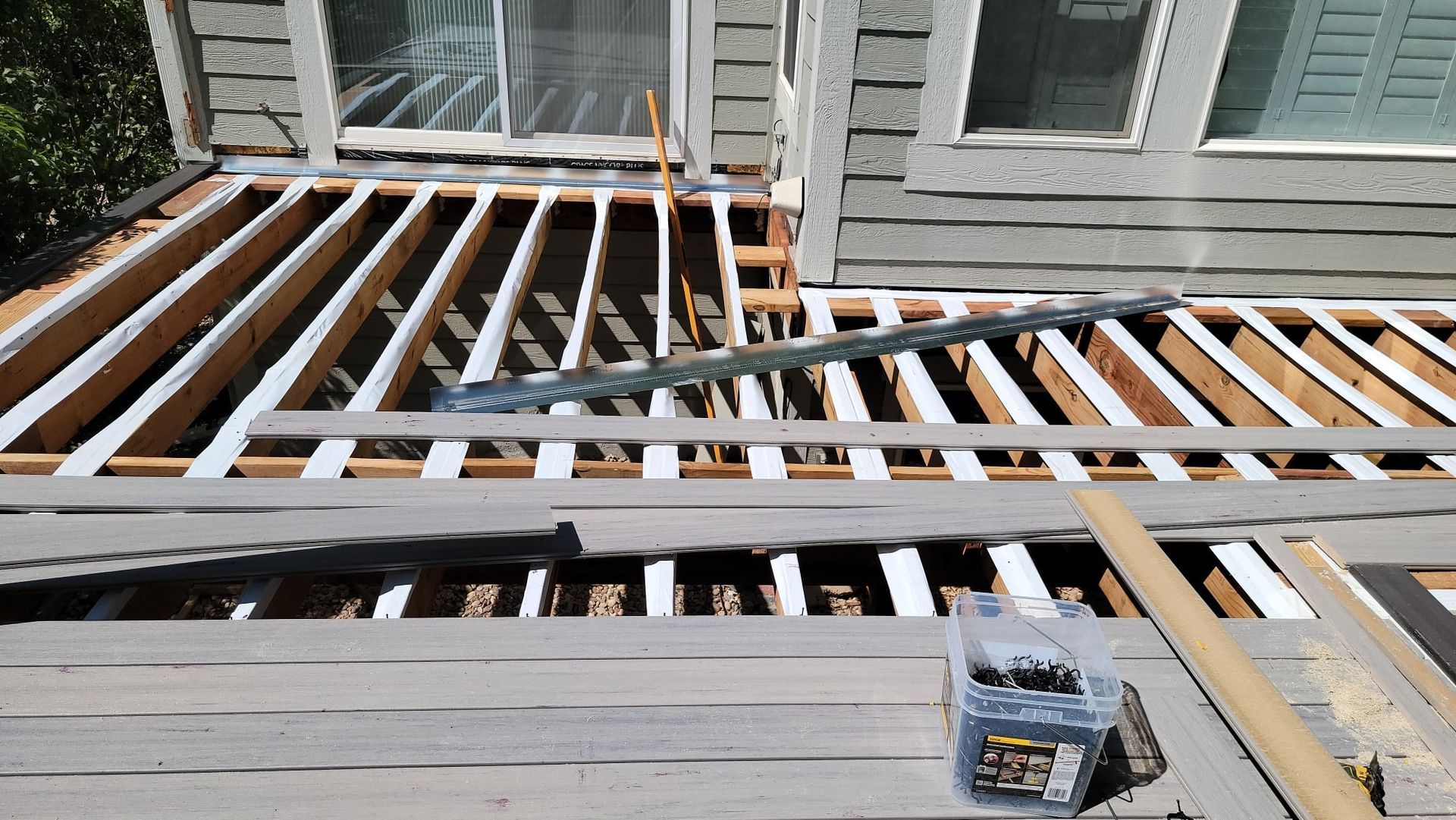 A wooden deck under construction with exposed floor joists, gray composite boards, and a box of fasteners nearby.