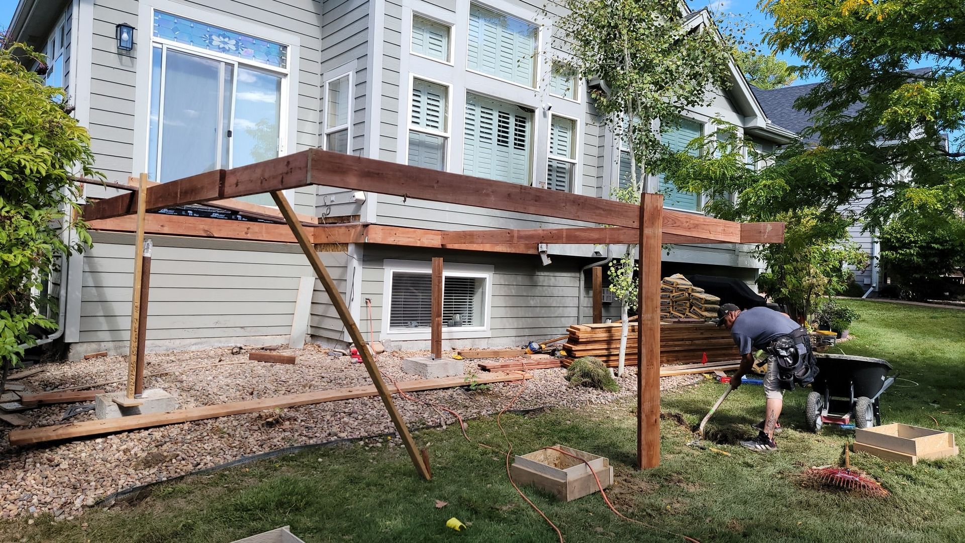 A person works in a backyard on the wooden frame of a deck being built against a two-story gray house.