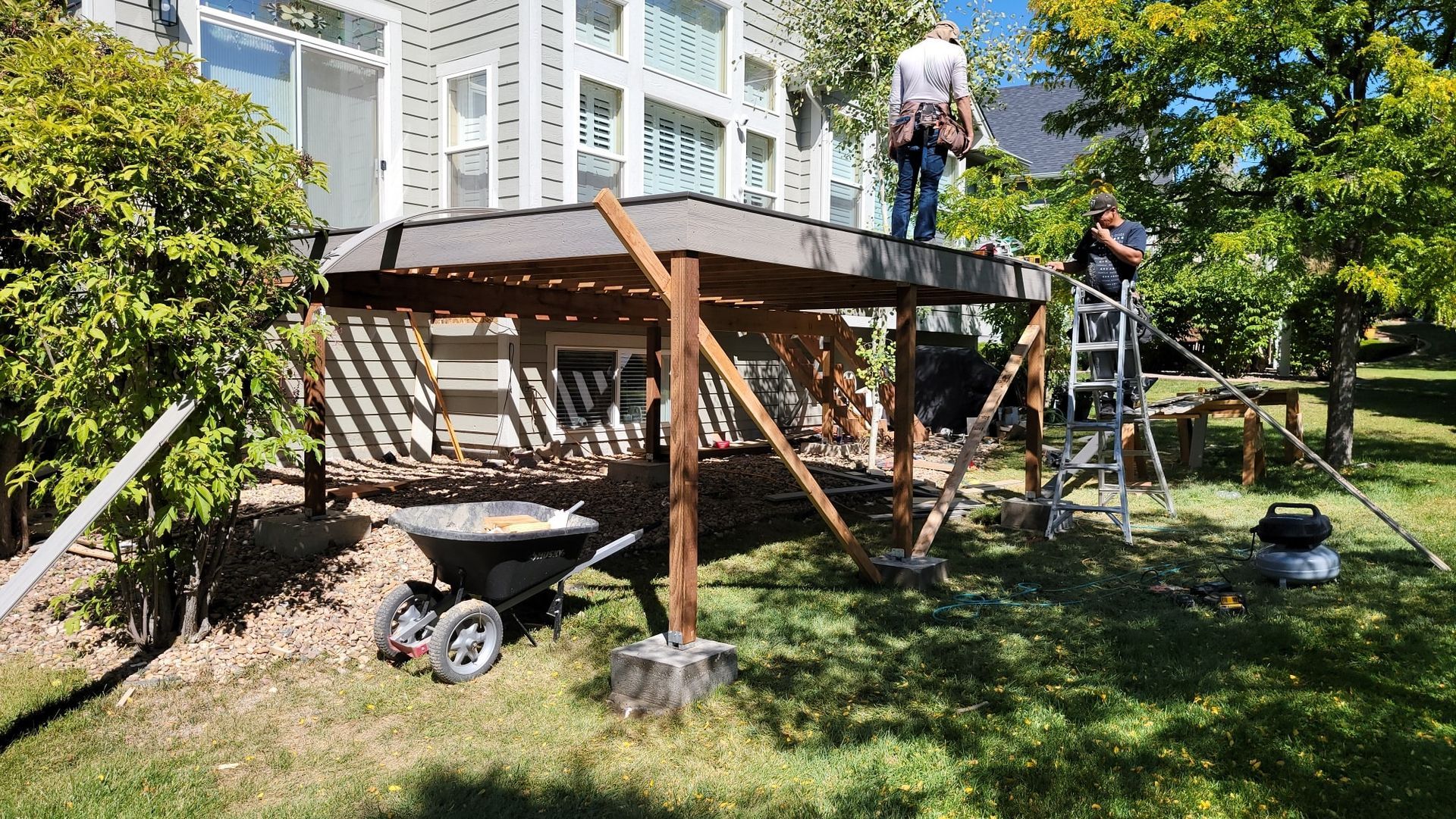 Two workers are building a wooden deck behind a house, with a ladder and wheelbarrow on the lawn nearby.