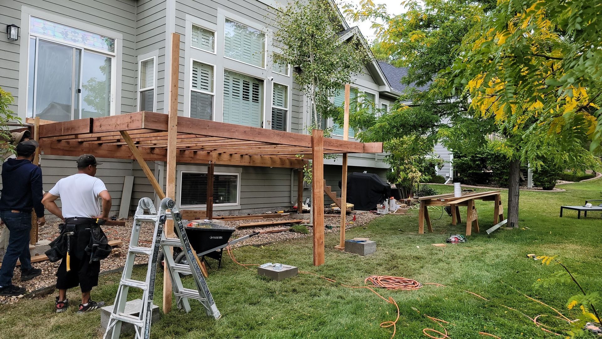 Two construction workers build a wooden deck frame attached to the back of a house in a residential backyard.