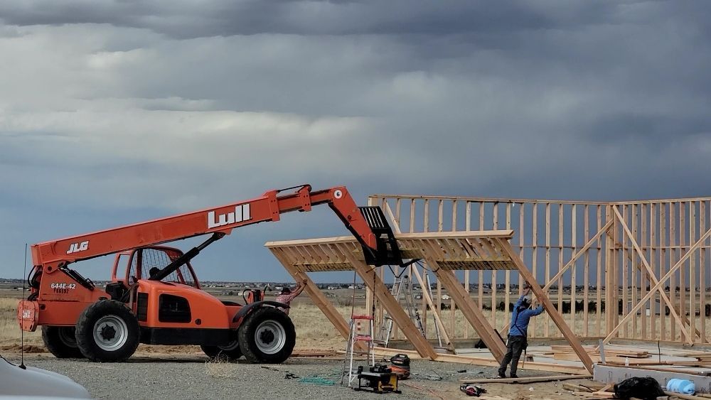 An orange telescopic forklift lifts a wooden wall frame into place at a construction site under a cloudy sky.