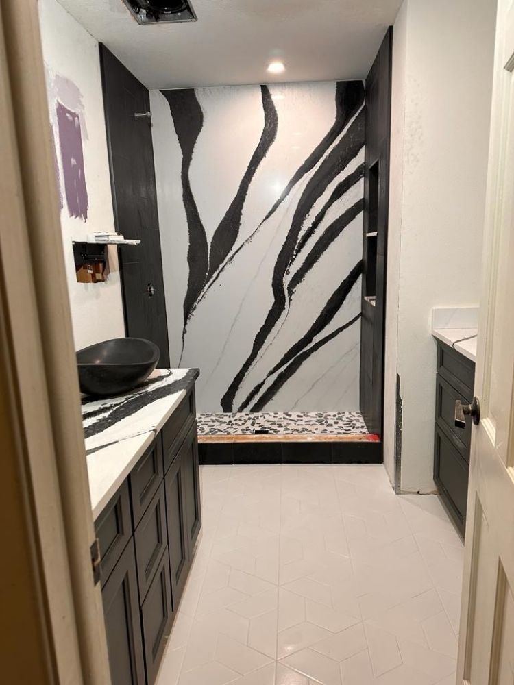 A bathroom with dark grey cabinets, white countertops, a black vessel sink, and a shower with bold black and white veining.
