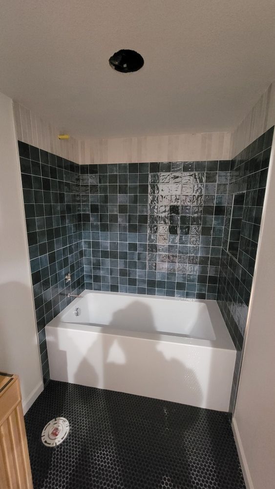 A white bathtub is surrounded by dark, square-tiled walls and features a black hexagon-tiled floor in a bathroom.