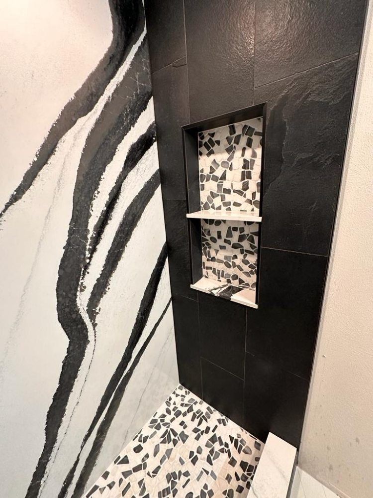 A shower corner featuring black textured wall tiles, a black-framed niche with patterned tiles, and a terrazzo floor.