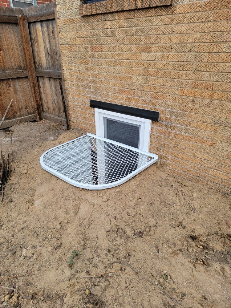 A white metal window well grate sits over a basement window against a brick exterior wall near a wooden fence.