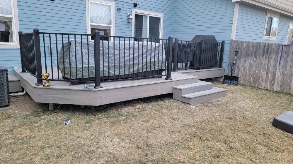 A gray composite deck with black railings and stairs against the side of a light blue house in a yard.