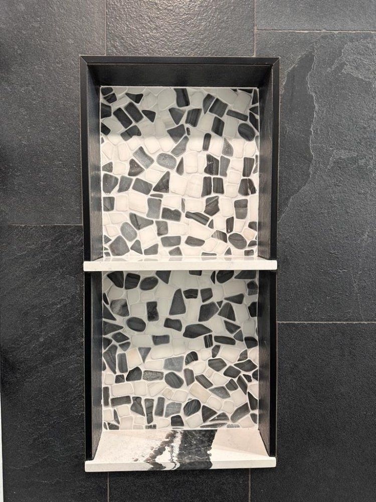 A black-framed shower niche with two shelves and a black-and-white terrazzo-style tile back, set in a dark tile wall.