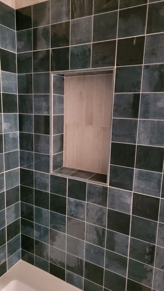 A shower wall with dark blue-grey square tiles and an unfinished, light wood-toned recessed shelf in the center.