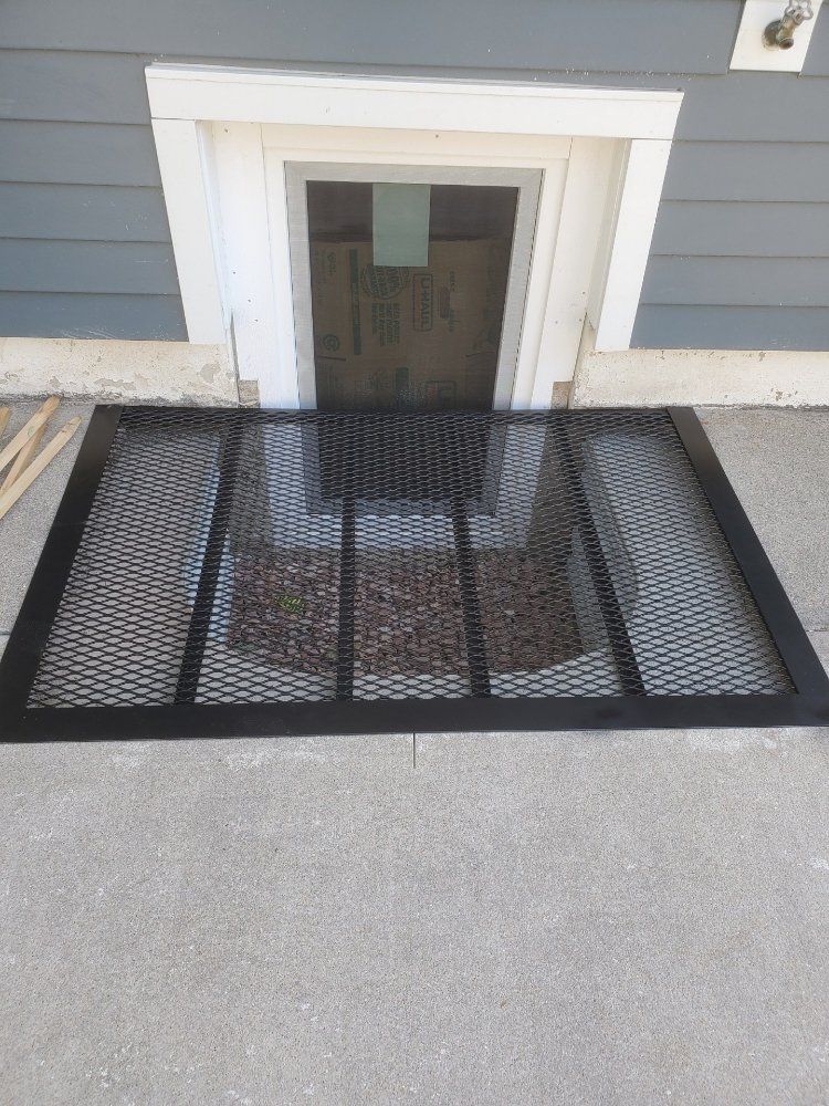 A black metal safety grate covers a window well against the gray siding of a house foundation.