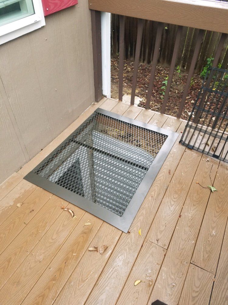 A metal-grated egress window well embedded in a wooden deck surface next to a house wall.