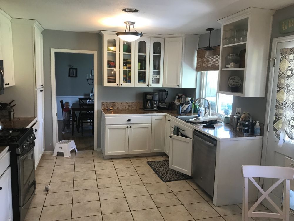 A brightly lit kitchen with white cabinets, tile floors, and stainless steel appliances, opening into a dining room.