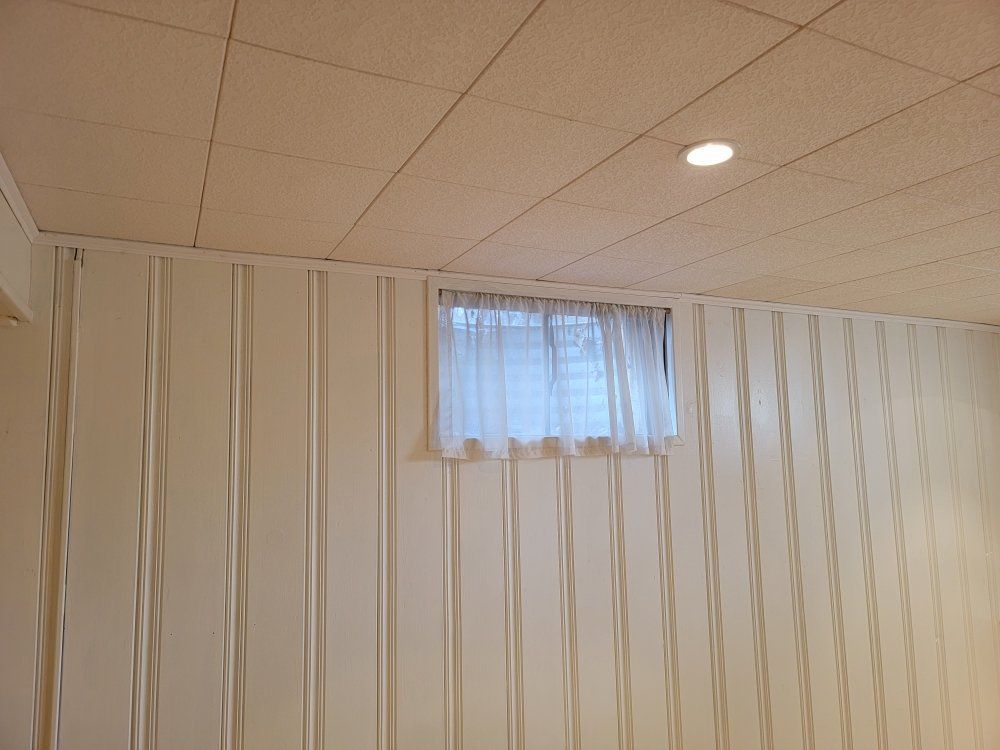 A basement wall covered in vertical beige paneling with a small window featuring white curtains and a tiled ceiling.