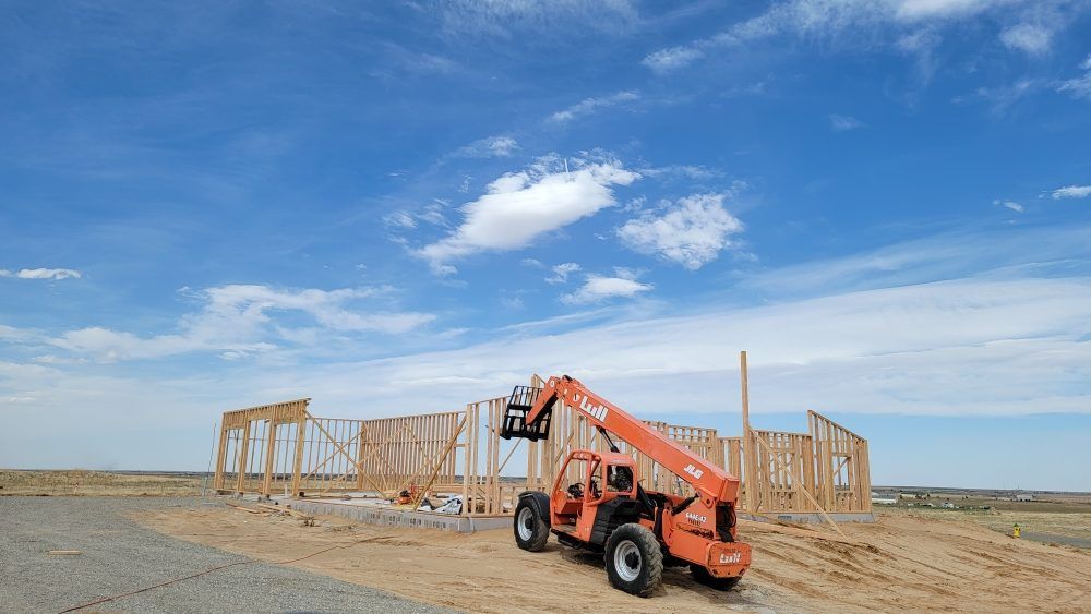 An orange telehandler parked on a dirt lot next to the wooden frame of a building under a bright blue sky.