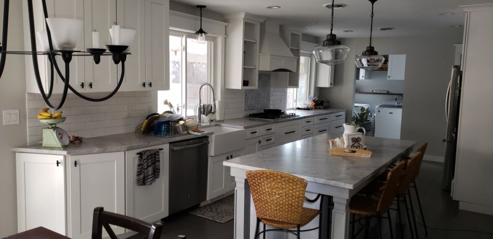 A bright kitchen with white cabinets, a central island with wicker stools, and a large chandelier over the countertop.