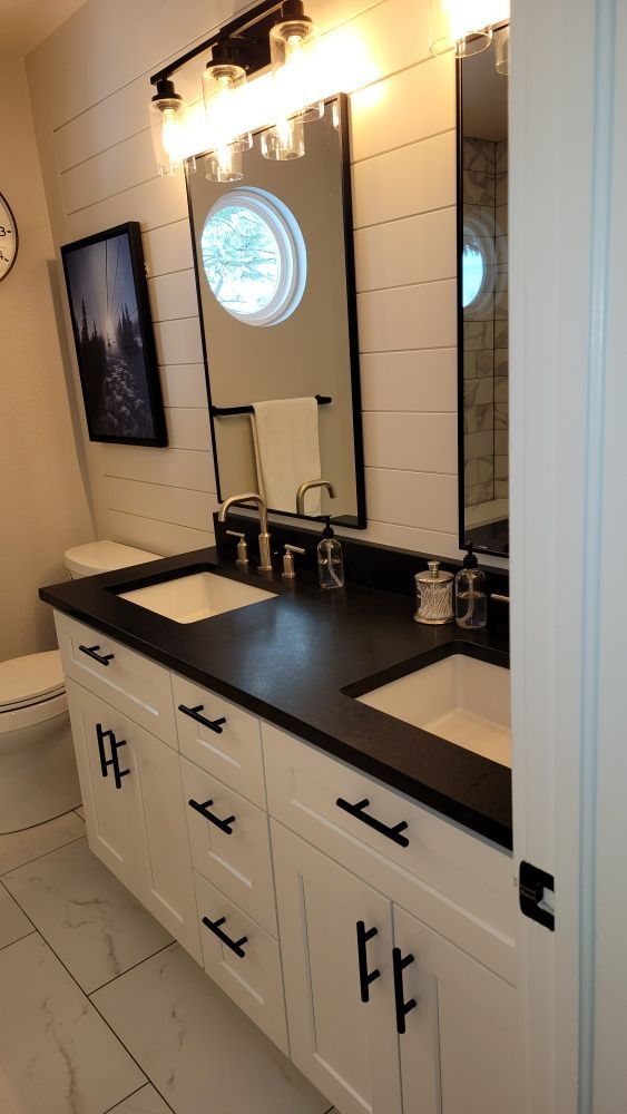 Modern white bathroom vanity with black hardware, dark countertops, dual sinks, shiplap walls, and two framed mirrors.
