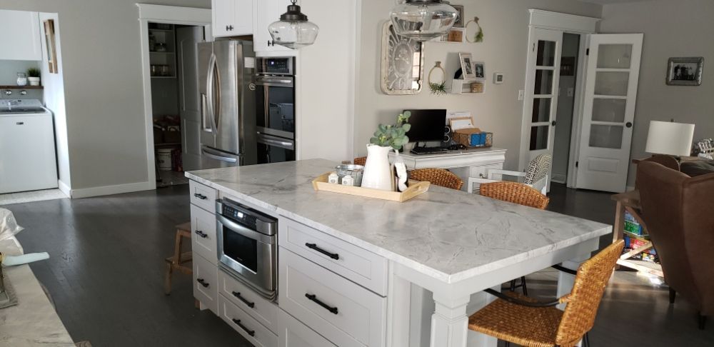 A modern kitchen features a large white island with a marble countertop, a built-in microwave, and wicker stools.