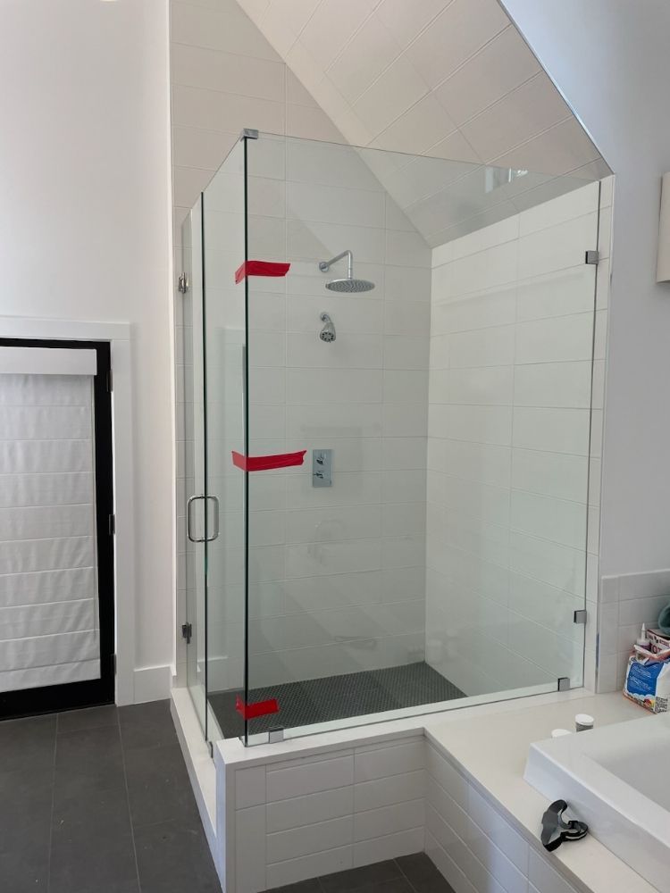 A frameless glass shower enclosure sits on a tiled base in a modern bathroom, with red tape marking the glass joints.