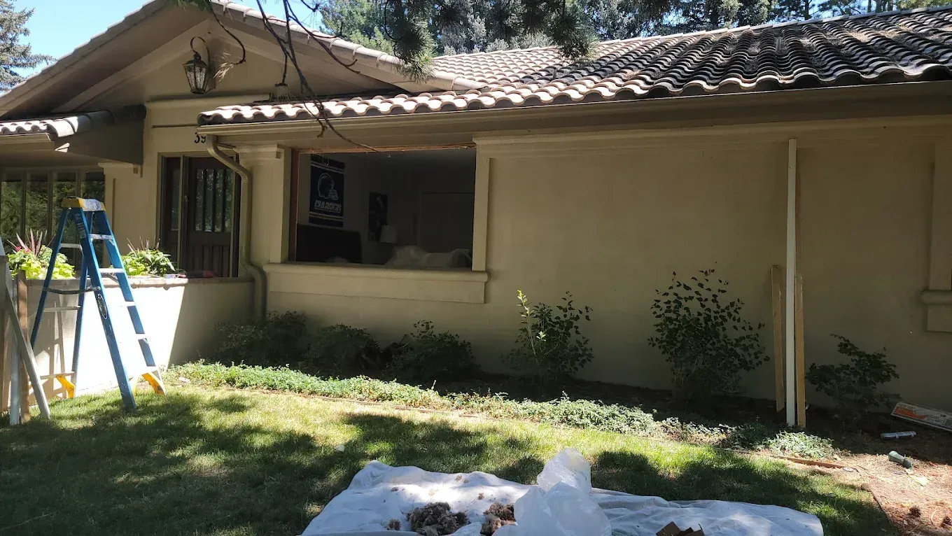Exterior of a tan house during a renovation, featuring an open window frame, a step ladder, and landscaping.