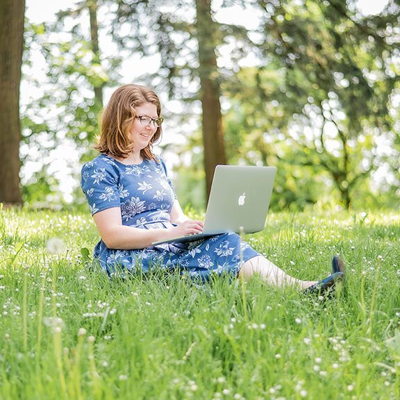 Renee sitting in a sunny, daisy-filled field with her laptop on her lap.