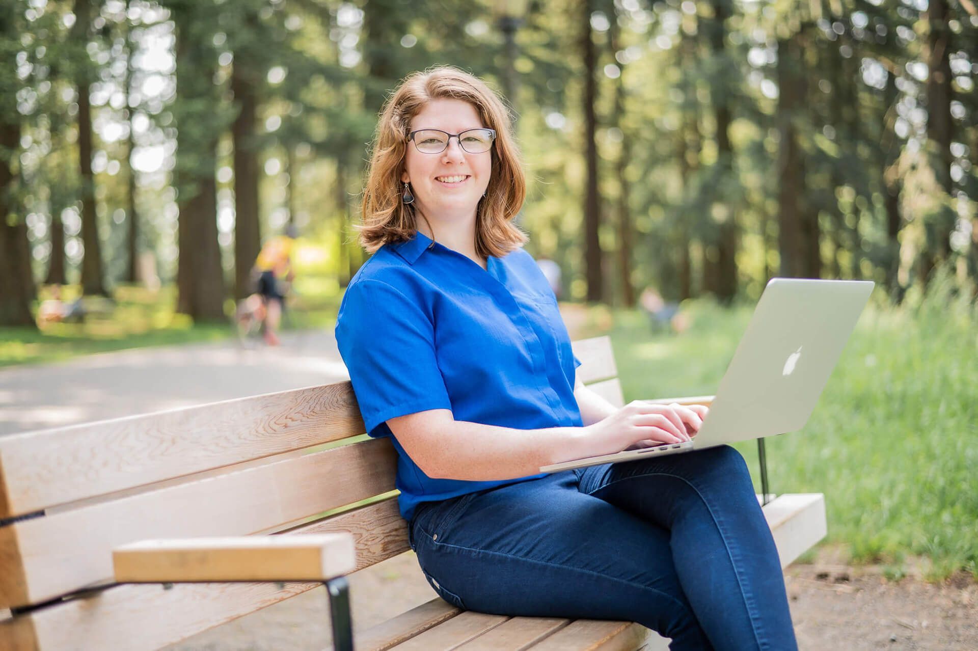 Photo of Renee, with shoulder length red hair and glasses. Smiling and sitting on a wooden bench in a park with her laptop.