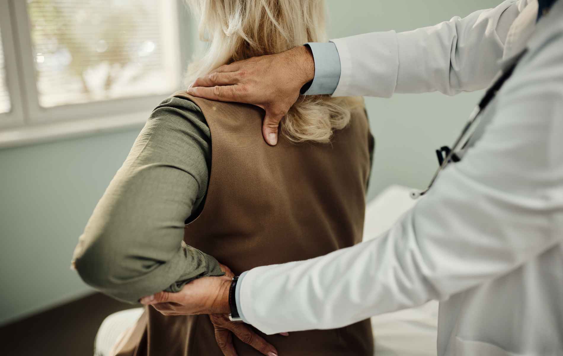 Doctor examining a patient's back in an office, hands on back, wearing a white coat.
