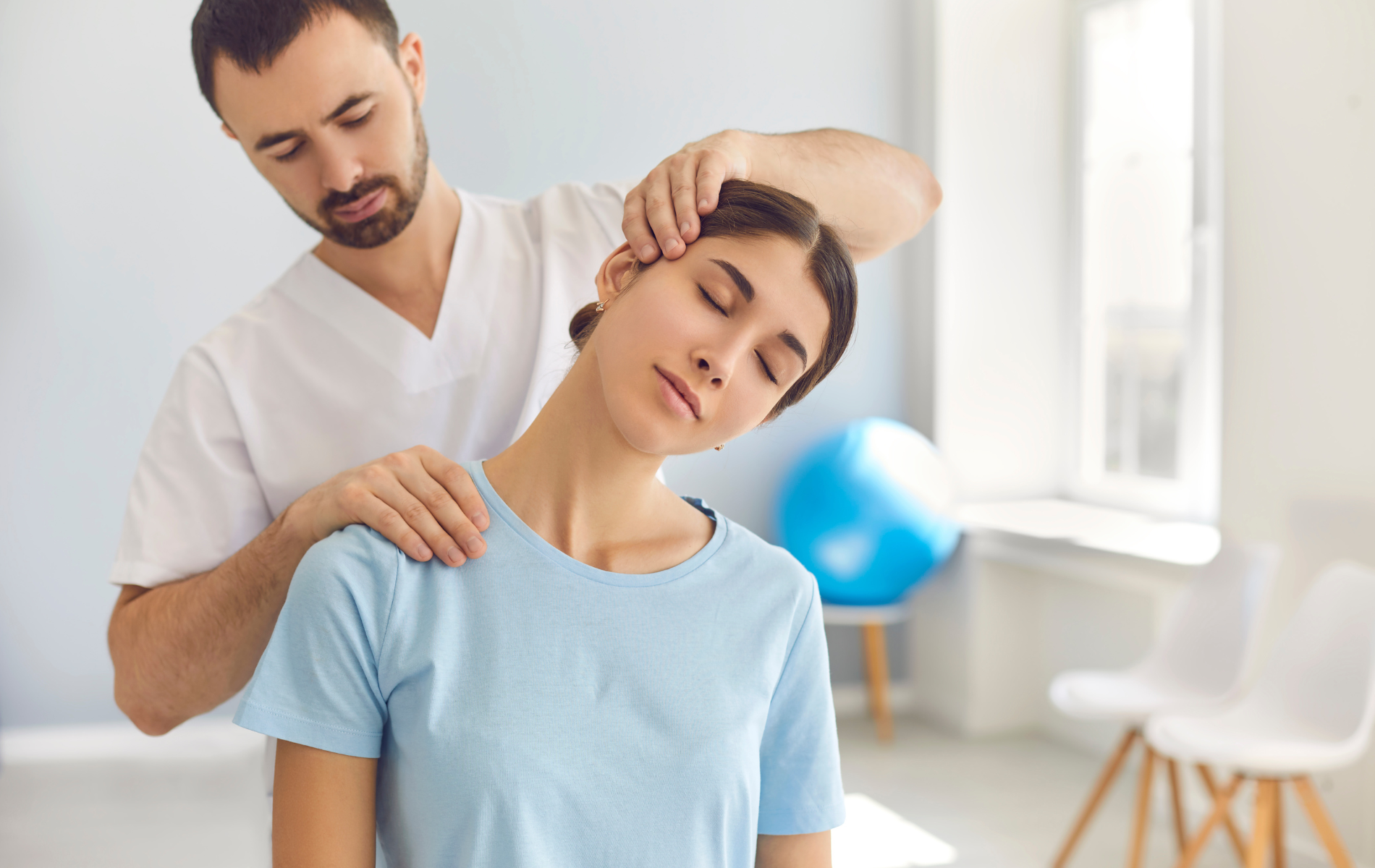 Man adjusting woman's neck in a light-filled room with exercise ball and chairs. Woman's eyes are closed.