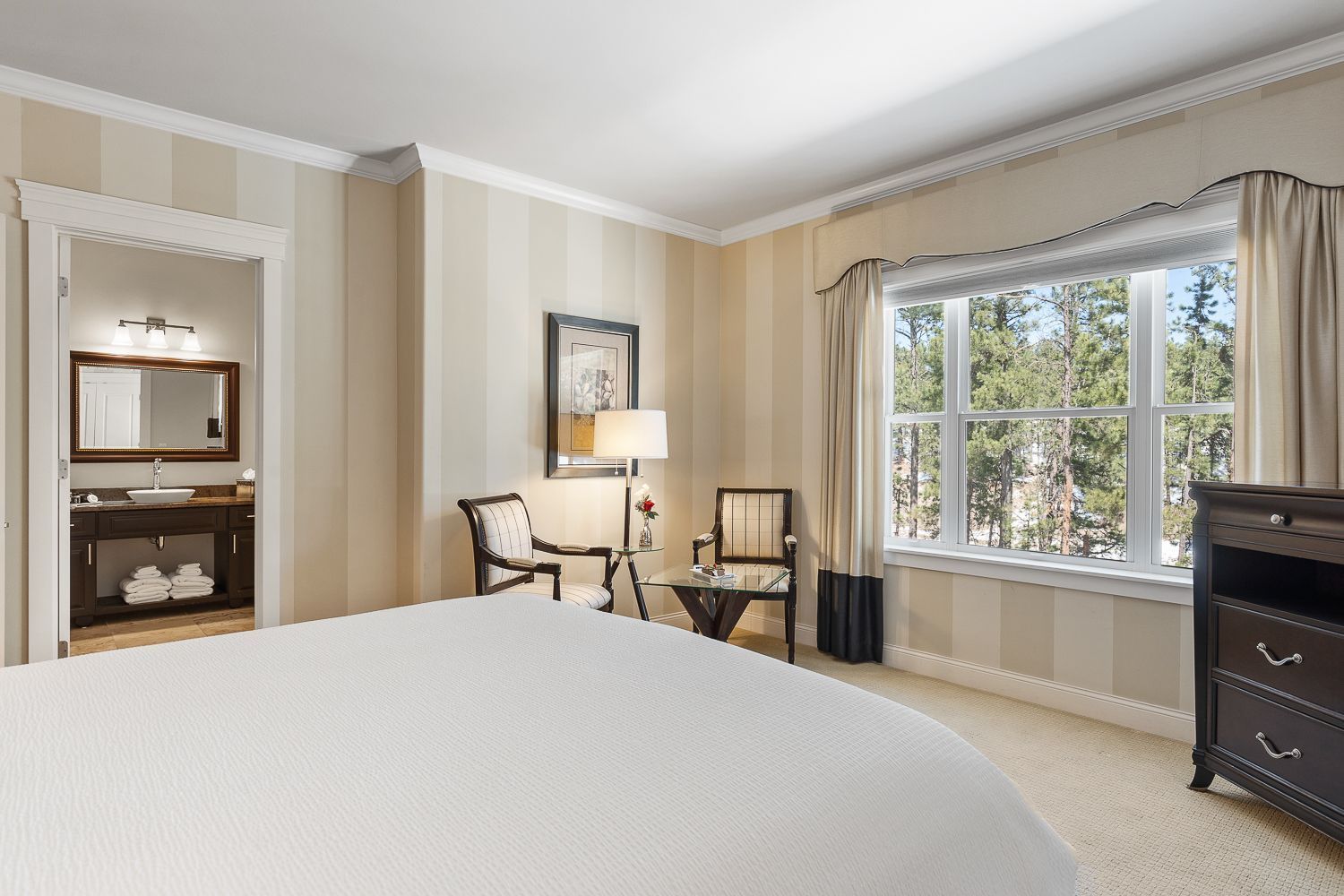 A hotel room with a king size bed , chair , dresser and window.