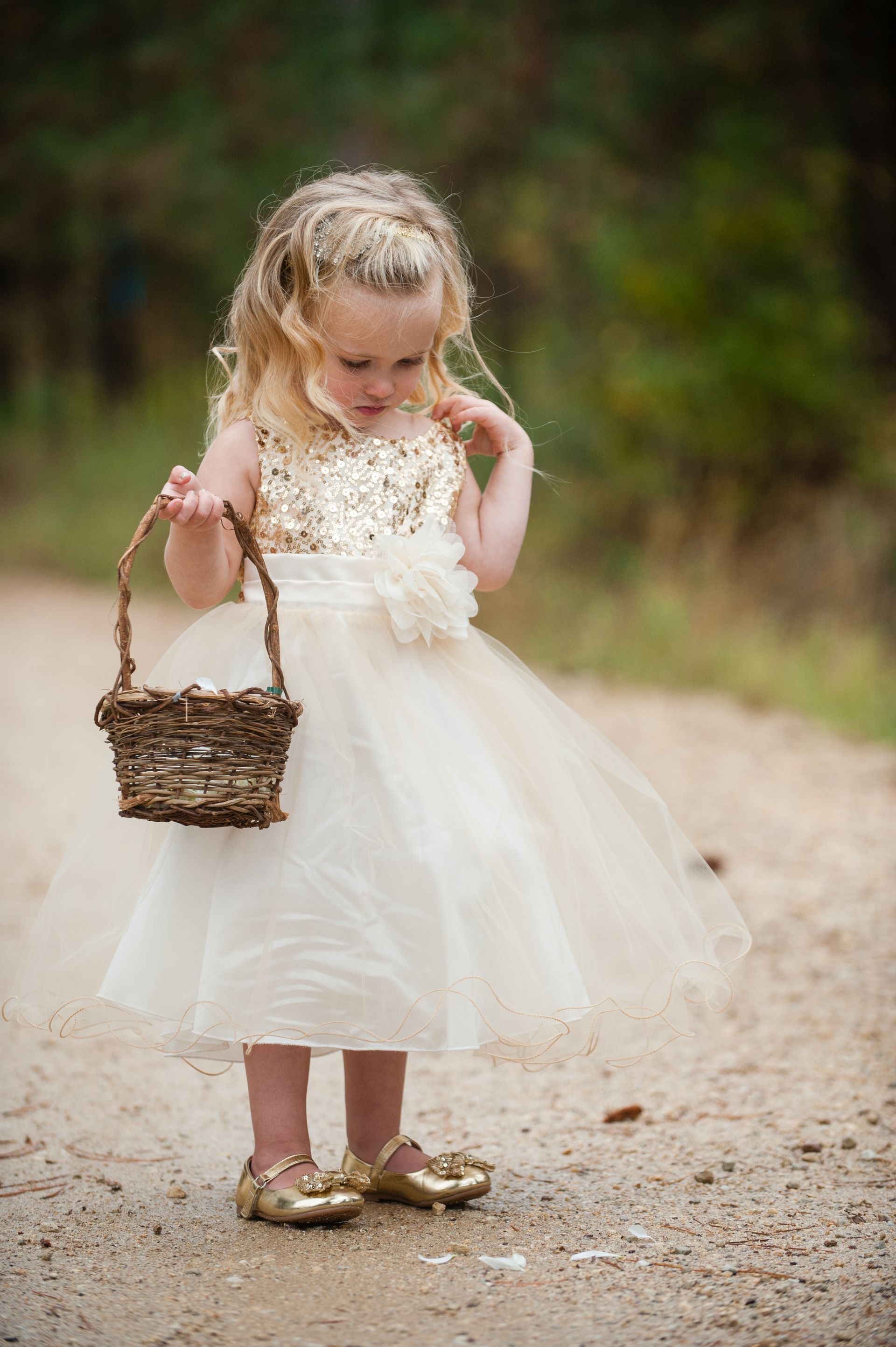 A flower girl in a white dress is holding a basket on a dirt road.