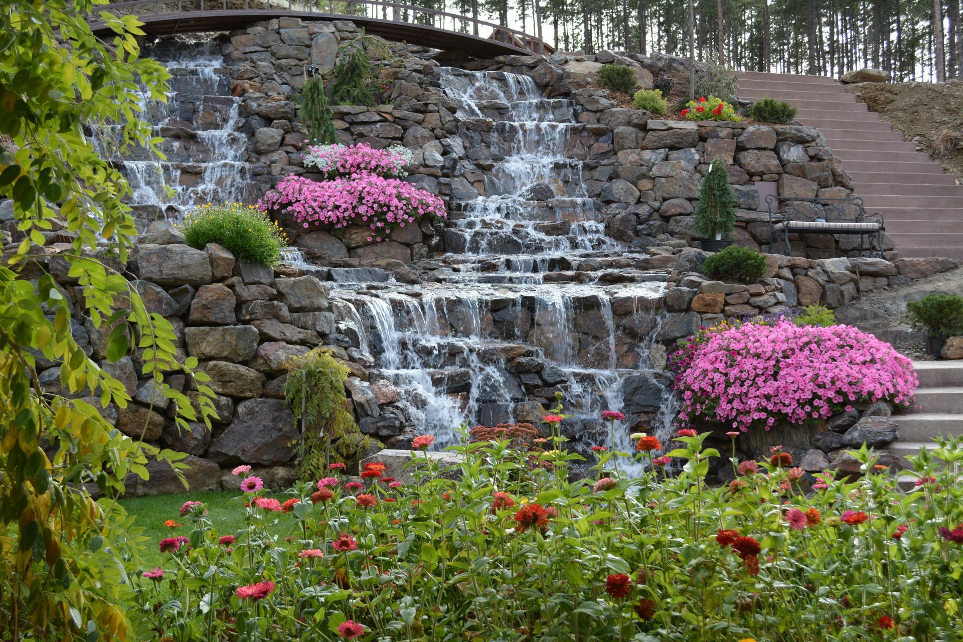 There is a waterfall in the middle of a garden surrounded by flowers.
