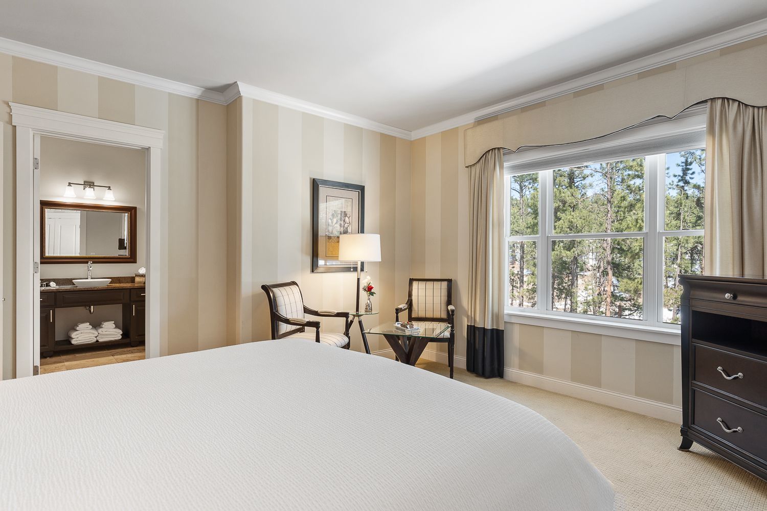 A hotel room with a king size bed , chair , dresser and window.