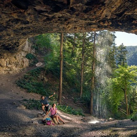 A man and a woman are standing under a cave near a waterfall.