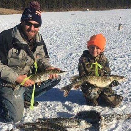 A man and a boy are kneeling in the snow holding fish.