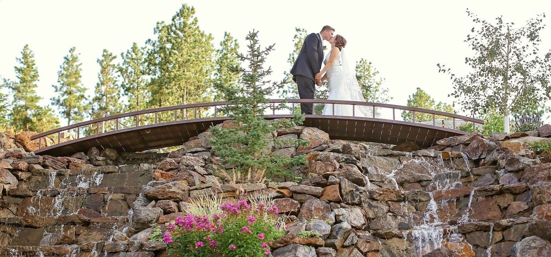 A bride and groom are kissing on a bridge over a waterfall.
