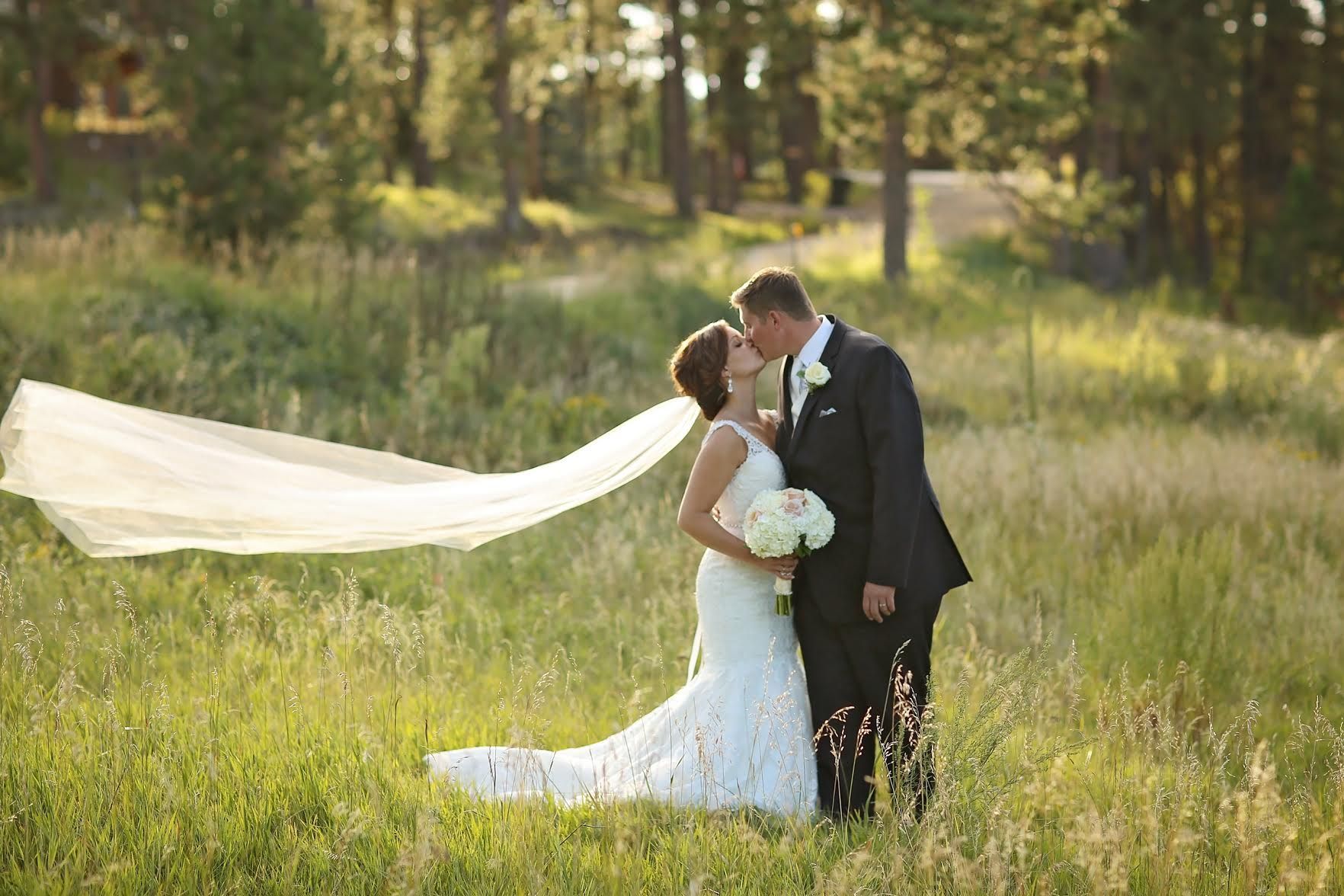 A bride and groom are kissing in a field with a veil blowing in the wind.