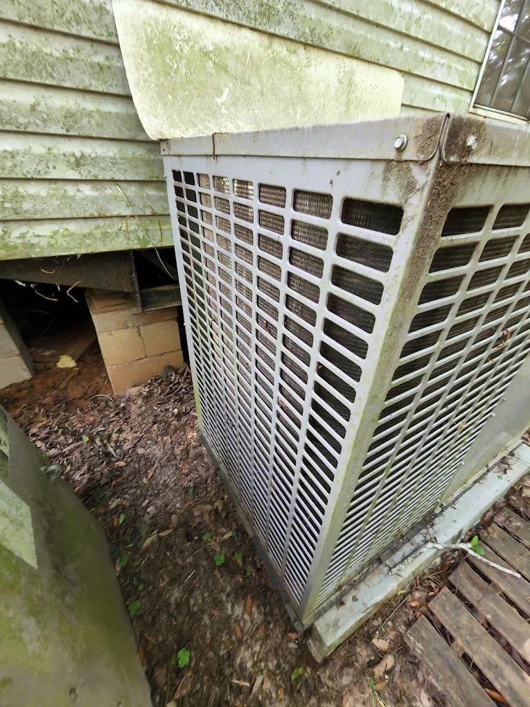 Air conditioner unit on the side of a house, with a weathered exterior and a wooden deck.