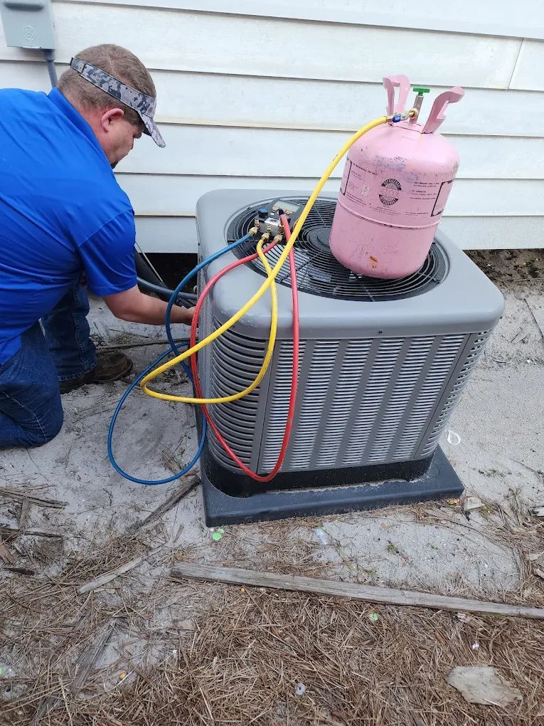 Man repairs AC unit, connecting hoses from pink gas tank. Outdoors, near house siding.