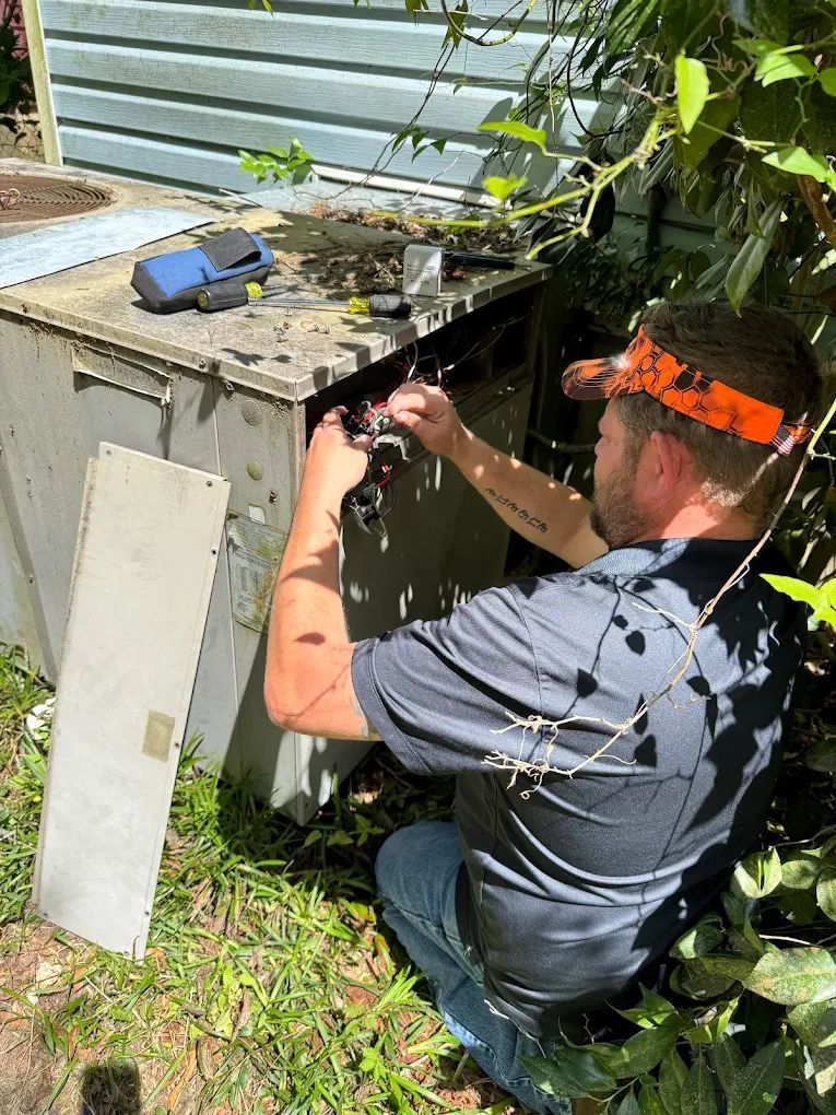Man in orange visor repairs AC unit outdoors, holding wires, surrounded by grass and foliage.
