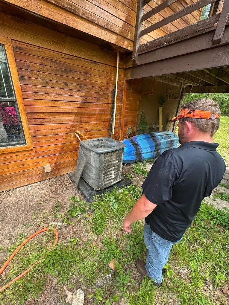 Man examines air conditioner unit next to wood cabin. He wears a hat and jeans. Green grass surrounds.