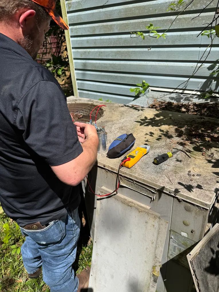 Man in jeans and black shirt tests an AC unit with a multimeter outdoors.
